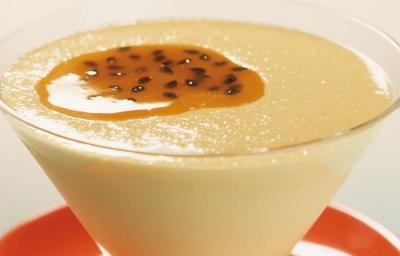 Fotografia em tons de amarelo e vermelho de uma bancada azul e branca com um pratinho vermelho e sobre ele uma taça com o creme de maracujá decorado com polpa e sementes de maracujá.