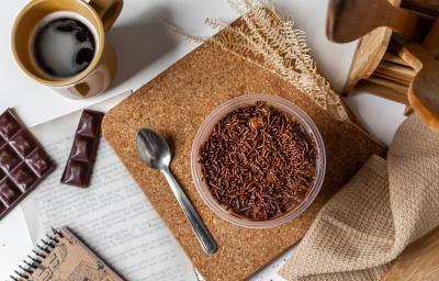 Fotografia em tons de madeira com um pote de plástico ao centro. Dentro do pote existe uma receita vista de cima de um bolo de pole de cenoura com cobertura de brigadeiro e granulados