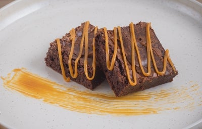 Foto aproximada de duas fatias cortadas da receita de brownie clássico de chocolate, decoradas com fios de doce de leite feito com Leite Moça cozido Foto aproximada de duas fatias cortadas da receita de brownie clássico de chocolate, decoradas com fios de doce de leite feito com Leite Moça cozido