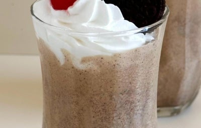 Fotografia em tons claros de dois copos de vidro com milkshake de negresco, decorados com chantilly, cereja e um biscoito Negresco inteiro.