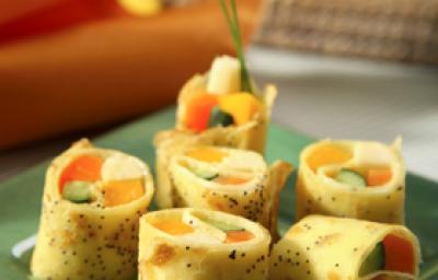 Fotografia em tons de laranja, amarelo e verde, com pedaços de panqueca recheadas com legumes sobre peça de vidro em tom verde, ao lado guardanapo laranja, ao fundo guardanapos de cores variadas, tudo sobre bancada em tom cinza.