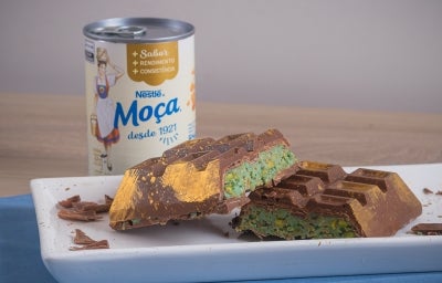 Barra de Dubai, receita de uma barra de chocolate recheada com um creme de pistache e macarrão, decorada com pó dourado e servida em um prato branco ao lado de uma lata de Leite Moça
