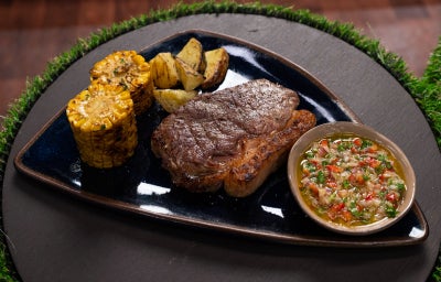 Bife de chorizo grelhado com milho e batatas, servido com chimichurri caseiro em cumbuca