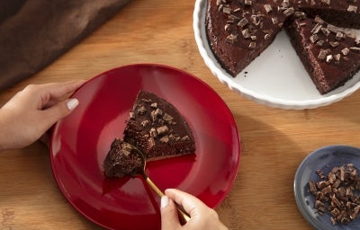 Fotografia de uma mão feminina segurando um prato vermelho raso, sobre uma mesa de madeira. No prato tem uma fatia de torta mousse de nescau extra cacau, ao lado tem um apoio branco com o restante da sobremesa.