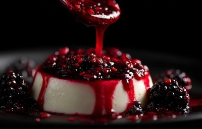 Fotografia em tons escuros de uma sobremesa com mousse com uma calda de frutas vermelhas sobre e ao redor da mousse, sobre um prato raso preto. Tem um pouco de calda caindo na mousse de uma colher de sopa.