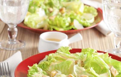 Fotografia em tons de vermelho e verde em uma bancada de madeira clara com um prato vermelho ao centro e a salada caesar com croutons em cima dele.