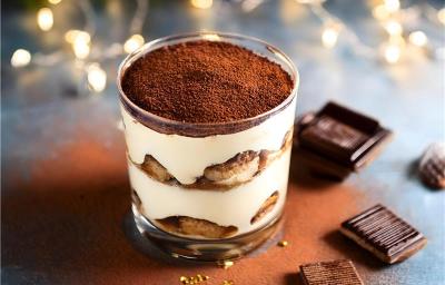 Fotografia de um copo de vidro pequeno com tiramisú. O copo está sobre uma mesa escura com cacau em pó polvilhado e alguns pedaços de chocolate. Fotografia de um copo de vidro pequeno com tiramisú. O copo está sobre uma mesa escura com cacau em pó polvilhado e alguns pedaços de chocolate.