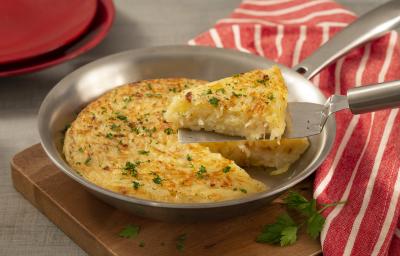Fotografia em tons de marrom, vermelho e cinza de uma tabua de madeira, uma frigideira de inox com a batata rosti e uma espátula retirando uma fatia. Ao lado um paninho listrado vermelho e branco, ao fundo dois pratos vermelhos. Fotografia em tons de marrom, vermelho e cinza de uma tabua de madeira, uma frigideira de inox com a batata rosti e uma espátula retirando uma fatia. Ao lado um paninho listrado vermelho e branco, ao fundo dois pratos vermelhos.