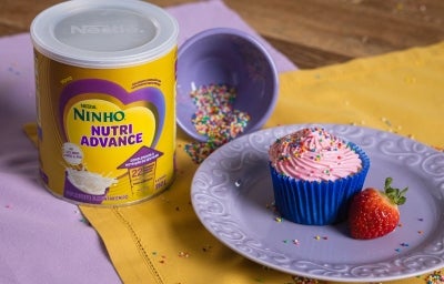 Fotografia de dois panos de mesa, roxo e amarelo, um sobre o outro em uma mesa de madeira. Sobre eles tem uma lata de NINHO Nutri Advance ao lado de um prato médio roxo com um cupcake e um morango inteiro, ao lado tem um potinho roxo com granulados. Fotografia de dois panos de mesa, roxo e amarelo, um sobre o outro em uma mesa de madeira. Sobre eles tem uma lata de NINHO Nutri Advance ao lado de um prato médio roxo com um cupcake e um morango inteiro, ao lado tem um potinho roxo com granulados.