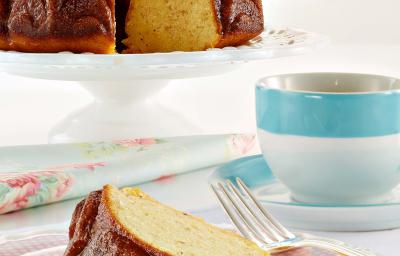 Fotografia em tons de azul em uma mesa branca com uma toalha branca, um prato de sobremesa com uma fatia de bolo de maracujá. Ao fundo, um suporte para bolo com o bolo inteiro e ao lado uma xícara com listra azul e chá dentro dela.