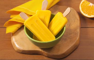 Fotografia em tons de laranja em uma bancada de madeira, uma tábua de madeira, um potinho verde e três picolés de manga com laranja dentro dele. Ao lado, um paninho amarelo e uma laranja partida ao meio.