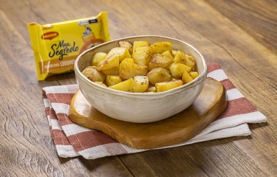 Fotografia em tons de amarelo e marrom de uma bancada de madeira com um pano retorcido amarelo, uma travessa branca oval com vários cubos de batatas dentro e salsinha polvilhada.