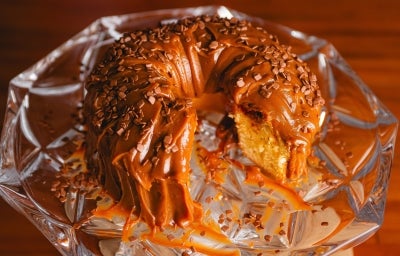 Fotografia em tons de marrom com um bolo de furo no meio ao centro. O bolo possui massa branca, frutas vermelhas e cobertura de brigadeiro de caramelo salgado.