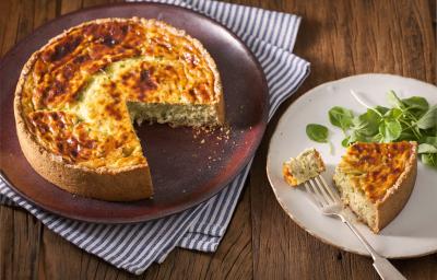 Fotografia em tons de marrom e branco de uma bancada de madeira com um paninho branco e azul, sobre ele um prato redondo vinho com um quiche. Ao lado um prato branco redondo com uma fatia de quiche.