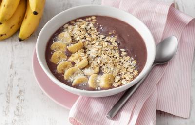 Fotografia em tons de branco e rosa de uma bancada de madeira branca com um paninho rosa, sobre ele um prato raso rosa com um recipiente redondo branco com o açaí. Ao lado uma colher e um cacho de banana.
