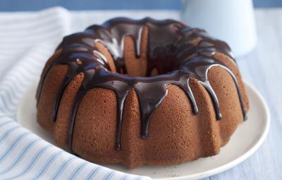 Fotografia em tons de azul em uma bancada de madeira clara com um pano branco listrado em azul, ao centro um prato branco raso com o bolo de chocolate, com cobertura de chocolate. Ao lado, um potinho com chocolate e uma jarra azul claro. Fotografia em tons de azul em uma bancada de madeira clara com um pano branco listrado em azul, ao centro um prato branco raso com o bolo de chocolate, com cobertura de chocolate. Ao lado, um potinho com chocolate e uma jarra azul claro.