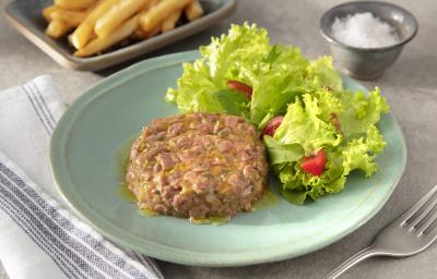 Fotografia em tons de verde em uma bancada de madeira clara, um pano azul, um prato verde claro com o steak tartare acompanhado de salada de alface e tomate cereja. Ao fundo, um recipiente com uma porção de batatas fritas.