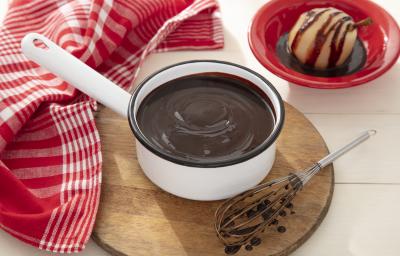 Fotografia em tons de vermelho de uma bancada, ao centro uma panela branca com uma calda de chocolate, em volta um pano vermelho e branco.