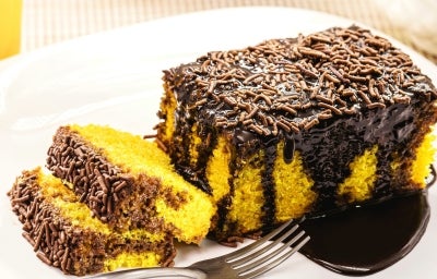 Um pedaço generoso de bolo de cenoura com cobertura de chocolate está em um prato branco. A cobertura é brilhante e farta, com granulado por cima. Ao lado, duas fatias menores destacam a massa amarelo-viva e fofinha, criando contraste com o chocolate Um pedaço generoso de bolo de cenoura com cobertura de chocolate está em um prato branco. A cobertura é brilhante e farta, com granulado por cima. Ao lado, duas fatias menores destacam a massa amarelo-viva e fofinha, criando contraste com o chocolate