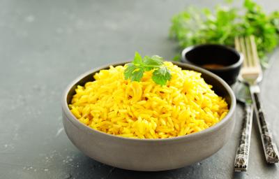 Fotografia em tons de verde com um prato fundo de cerâmica cinza ao centro. Em cima do prato existe um arroz com curry de cor amarelada. Por cima existe uma folha de salsinha.