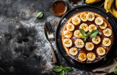 Bolo decorado com muitas fatias de banana sobre uma camada de calda de caramelo. Há uma folha de hortelã no centro para enfeitar. O bolo está em um prato preto, cercado por bananas maduras, talheres e açúcar de confeiteiro espalhado pela mesa.