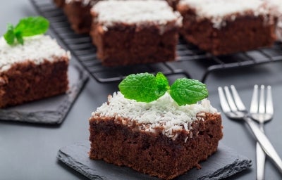 Fotografia de um quadrado de bolo de chocolate, coberto com coco ralado branco e decorado com uma folha de hortelã. O bolo está em uma tábua de ardósia escura, com mais pedaços ao fundo.