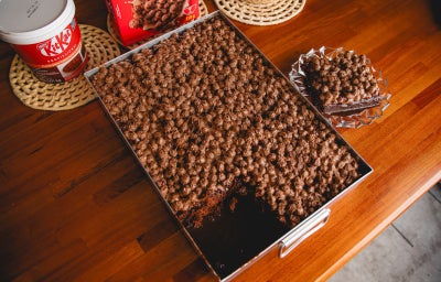 Fotografia mostra Bolo de chocolate úmido coberto com uma generosa camada de Pasta KITKAT® misturada ao NESCAU® Cereal, com brilho e textura crocante.