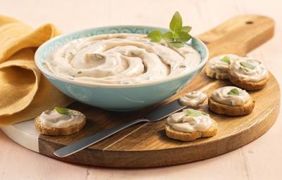 Foto em tons de bege com a receita de creme de ricota em um bowl azul claro em cima de uma tábua de madeira com 6 torradas com o creme em cima, além de uma faca prateada em frente ao bowl. Ao fundo um pano amarelo de decoração, isso numa mesa de madeirair