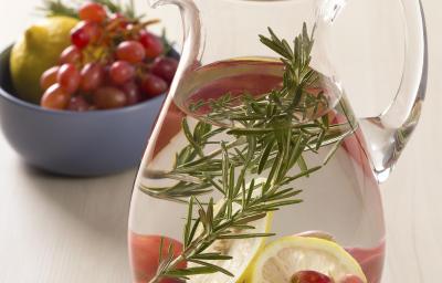 Fotografia em tons de branco, verde e roxo de uma bancada de madeira clara, ao centro uma jarra de vidro transparente com a água, limão, uva e o alecrim. Ao fundo em um recipiente azul as mesmas frutas estão dentro para decoração. Fotografia em tons de branco, verde e roxo de uma bancada de madeira clara, ao centro uma jarra de vidro transparente com a água, limão, uva e o alecrim. Ao fundo em um recipiente azul as mesmas frutas estão dentro para decoração.