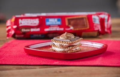 Fotografia de um tiramissu em camadas de creme e choco biscuit sobre um prato quadrado vermelho e raso com cacau em pó polvilhado. O prato está apoiado em um pano vermelho, e ao fundo, tem um pacote de choco biscuit.