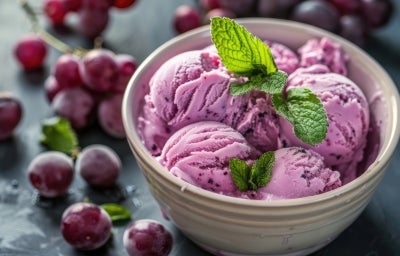 Fotografia em tons de roxo com um pote de sorvete ao centro. Dentro do pote existe uma receita de sorvete de uva de cor roxa finalizado com ramos de hortelã