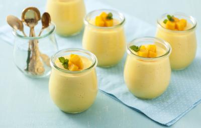 Fotografia em tons de azul e amarelo de uma bancada azul com um paninho azul com bolinhas brancas, um pote transparente com quatro colheres douradas e cinco potes transparentes com a mousse de manga, decorados com cubinhos de manga e uma folha de hortelã.