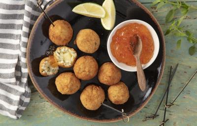 Fotografia em tons de verde em uma bancada de madeira verde, um pano listrado azul e cinza, um prato redondo preto com borda marrom e vários bolinhos de risoto em cima dele e um potinho branco com a geleia de pimenta para acompanhar. Fotografia em tons de verde em uma bancada de madeira verde, um pano listrado azul e cinza, um prato redondo preto com borda marrom e vários bolinhos de risoto em cima dele e um potinho branco com a geleia de pimenta para acompanhar.