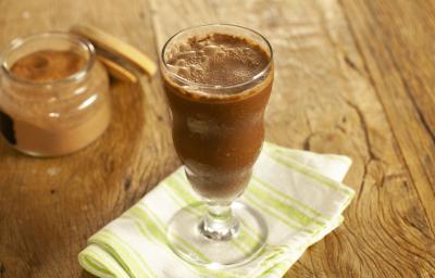 Fotografia em tons de marrom e branco de uma bancada de madeira com um paninho branco e verde, sobre ele um copo de vidro com o smoothie. Ao fundo um recipiente de vidro com cacau em pó. Fotografia em tons de marrom e branco de uma bancada de madeira com um paninho branco e verde, sobre ele um copo de vidro com o smoothie. Ao fundo um recipiente de vidro com cacau em pó.