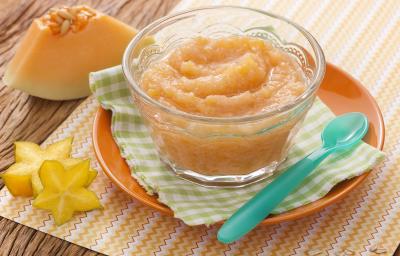 Fotografia em tons de laranja em uma bancada de madeira com um paninho xadrez verde e um potinho de vidro em cima com o mingauzinho de melão e carambola. Ao lado, um pedaço de um de melão e duas fatias cortadas de carambola. Fotografia em tons de laranja em uma bancada de madeira com um paninho xadrez verde e um potinho de vidro em cima com o mingauzinho de melão e carambola. Ao lado, um pedaço de um de melão e duas fatias cortadas de carambola.