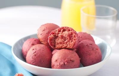 Fotografia em tons de azul e rosa em uma mesa branca com um pano azul ao lado e um prato branco fundo ao centro com os pães de beterraba. Ao fundo, uma jarra com suco de laranja e um copo de vidro. Fotografia em tons de azul e rosa em uma mesa branca com um pano azul ao lado e um prato branco fundo ao centro com os pães de beterraba. Ao fundo, uma jarra com suco de laranja e um copo de vidro.