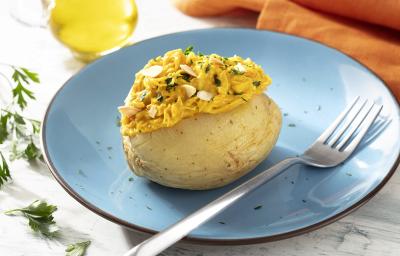 Fotografia em tons de amarelo em uma bancada de madeira de cor branca. Ao centro, um prato azul contendo a batata com um garfo ao lado. Ao fundo, há um pano laranja, um pote de azeite e algumas ervas espalhadas. Fotografia em tons de amarelo em uma bancada de madeira de cor branca. Ao centro, um prato azul contendo a batata com um garfo ao lado. Ao fundo, há um pano laranja, um pote de azeite e algumas ervas espalhadas.