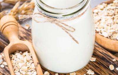 Fotografia em tons claros de um pote decorativo com bebida vegetal de coco sobre uma mini tábua de madeira escura retangular. Ao lado do pote tem aveia em flocos espalhadas e dentro de uma colher de madeira longa. Fotografia em tons claros de um pote decorativo com bebida vegetal de coco sobre uma mini tábua de madeira escura retangular. Ao lado do pote tem aveia em flocos espalhadas e dentro de uma colher de madeira longa.