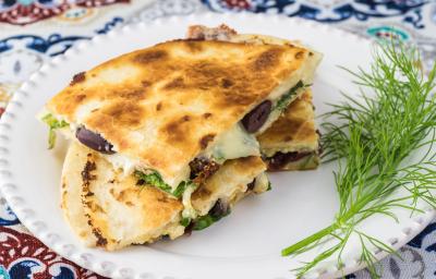 Fotografia em tons de branco com um prato branco ao centro. Em cima do prato existe uma receita de quesadilla de queijo com espinafre.