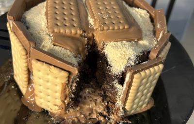 Fotografia de um bolo de chocolate com brigadeiro e farinha láctea polvilhada. Ao redor do bolo tem chocotrio de amendoim grudado. O bolo está sobre um prato de vidro preto raso. Fotografia de um bolo de chocolate com brigadeiro e farinha láctea polvilhada. Ao redor do bolo tem chocotrio de amendoim grudado. O bolo está sobre um prato de vidro preto raso.