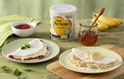 Fotografia em tons de verde de uma bancada verde com um prato redondo branco, sobre ele uma tapioca. Ao lado uma tábua de madeira com um prato redondo e uma tapioca. Ao fundo uma lata de ninho, um cacho de bananas e dois recipiente com mel e goiabada