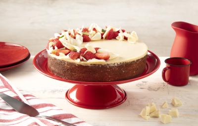 Fotografia em tons de vermelho em uma bancada bege com um paninho listrado branco e vermelho e ao centro um suporte vermelho com o bolo de três leites com ganache de chocolate branco. Ao lado, pedaços de chocolate branco, uma caneca e uma jarra vermelhas. Fotografia em tons de vermelho em uma bancada bege com um paninho listrado branco e vermelho e ao centro um suporte vermelho com o bolo de três leites com ganache de chocolate branco. Ao lado, pedaços de chocolate branco, uma caneca e uma jarra vermelhas.
