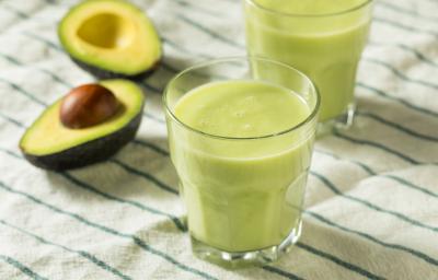 Fotografia de dois copos de vidro com vitamina de abacate. Ao fundo, ao lado do segundo copo, tem um abacate cortado ao meio, sobre uma toalha de mesa branca com listras verdes. Fotografia de dois copos de vidro com vitamina de abacate. Ao fundo, ao lado do segundo copo, tem um abacate cortado ao meio, sobre uma toalha de mesa branca com listras verdes.