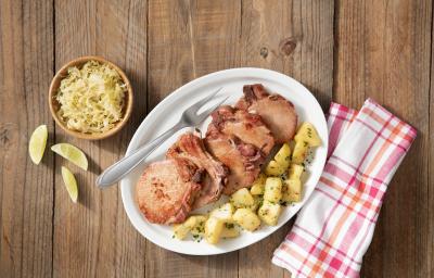 Fotografia em tons de branco de marrom, com bancada de madeira vista de cima, ao centro travessa branca contendo bisteca suína, batata picada e um garfo, no entorno potinho com repolho e fatias de limão e guardanapo quadriculado em branco e vermelho. Fotografia em tons de branco de marrom, com bancada de madeira vista de cima, ao centro travessa branca contendo bisteca suína, batata picada e um garfo, no entorno potinho com repolho e fatias de limão e guardanapo quadriculado em branco e vermelho.