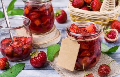 Potes de vidro com compota de morango, cercados por morangos frescos e cesta de frutas ao fundo.