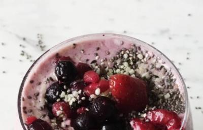 Fotografia em tons de vermelho em uma bancada de mármore branco, com um copo de vidro largo e grande com o smoothie de baunilha com frutas vermelhas dentro dele.