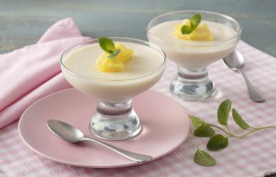 Fotografia em tons de rosa e amarelo de uma bancada, ao centro um paninho quadriculado rosa e branco, um prato rosa com uma taça com o mousse e pedacinhos de abacaxi e folha de hortelã. Ao lado uma taça igual e folhas de hortelã. Fotografia em tons de rosa e amarelo de uma bancada, ao centro um paninho quadriculado rosa e branco, um prato rosa com uma taça com o mousse e pedacinhos de abacaxi e folha de hortelã. Ao lado uma taça igual e folhas de hortelã.
