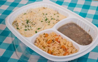 Foto aproximada de uma marmita branca de plástico com três divisões, uma com o frango com creme de milho, outra com arroz e outra com feijão. Ela está sobre um pano azul e branco quadriculado