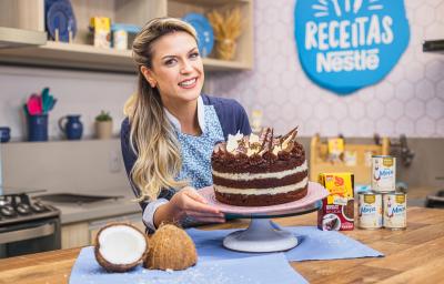 Imagem da confeiteira e influenciadora Beca Milano segurando a receita de bolo Naked de Prestígio atrás de uma bancada de cozinha, com pedaços de coco fresco e alguns produtos do lado direito Imagem da confeiteira e influenciadora Beca Milano segurando a receita de bolo Naked de Prestígio atrás de uma bancada de cozinha, com pedaços de coco fresco e alguns produtos do lado direito