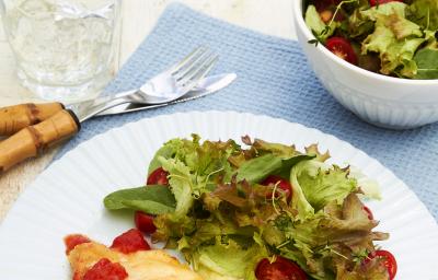 Fotografia em tons de azul em uma bancada de madeira clara, um pano azul, um prato branco redondo raso com o frango à parmegiana e uma salada verde em cima dele. Ao lado, talher, um copo com água e rodelas de limão e um potinho com mais salada.